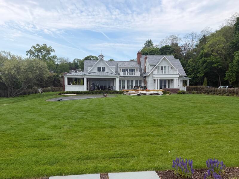 Rear exterior of a large shingle-style estate with manicured lawn, gabled roofs, and covered porches where motorized screens are being installed