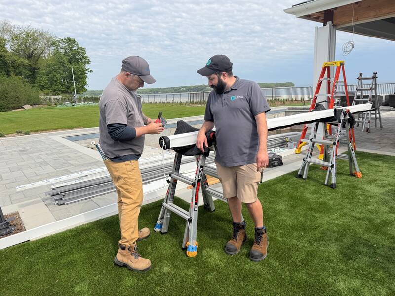 Installer and project lead inspecting a motorized screen tube and fabric on a ladder beside the reflecting pool