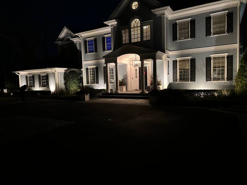 Shingle-style facade with warm uplighting at columns, gables, and portico